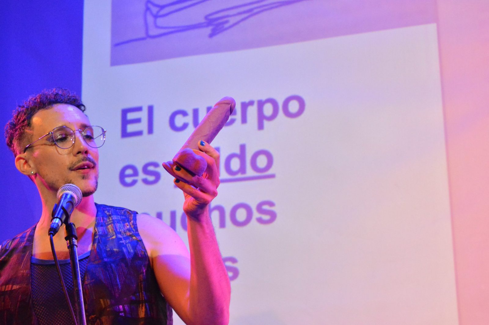Nicol López en el escenario con un dildo en la mano, frente a la proyección «El cuerpo es dildo»